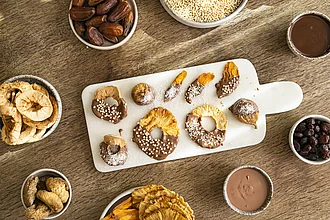 Rapunel Kuvertüre halbseitig auf Rapunzel Trockenfrüchten liegend auf einem hellen Brett. Rundherum stehen kleine Schüsseln mit Trockenfrüchten und flüssiger Schokolade