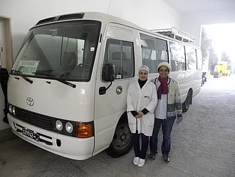 Samia Chibani steht mit einer Mitarbeiterin vor dem Werksbus auf dem ein HAND IN HAND-Logo abgebildet ist.