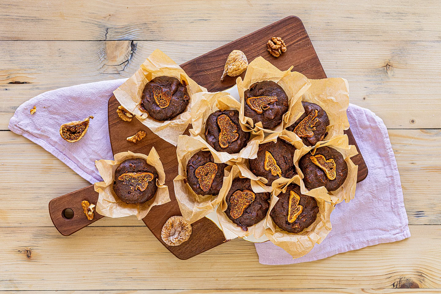 Schoko-Muffins mit Feigen In der Frontsicht sieht man mehrere Schoko-Muffins in Papierförmchen, die auf einem dunklen Holzbrett angerichtet sind. Jeder Muffin ist mit einer getrockneten Feigenscheibe garniert. Um die Muffins herum liegen halbierte getrocknete Feigen und Walnüsse. Unter den Holzbrettern ist ein fliederfarbenes Küchentuch drapiert.