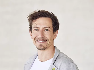 Rapunzel Geschäftsführer Leonhard Wilhelm im grauen Sakko schaut lächelnd in die Kamera und steht vor grauer Betonmauer