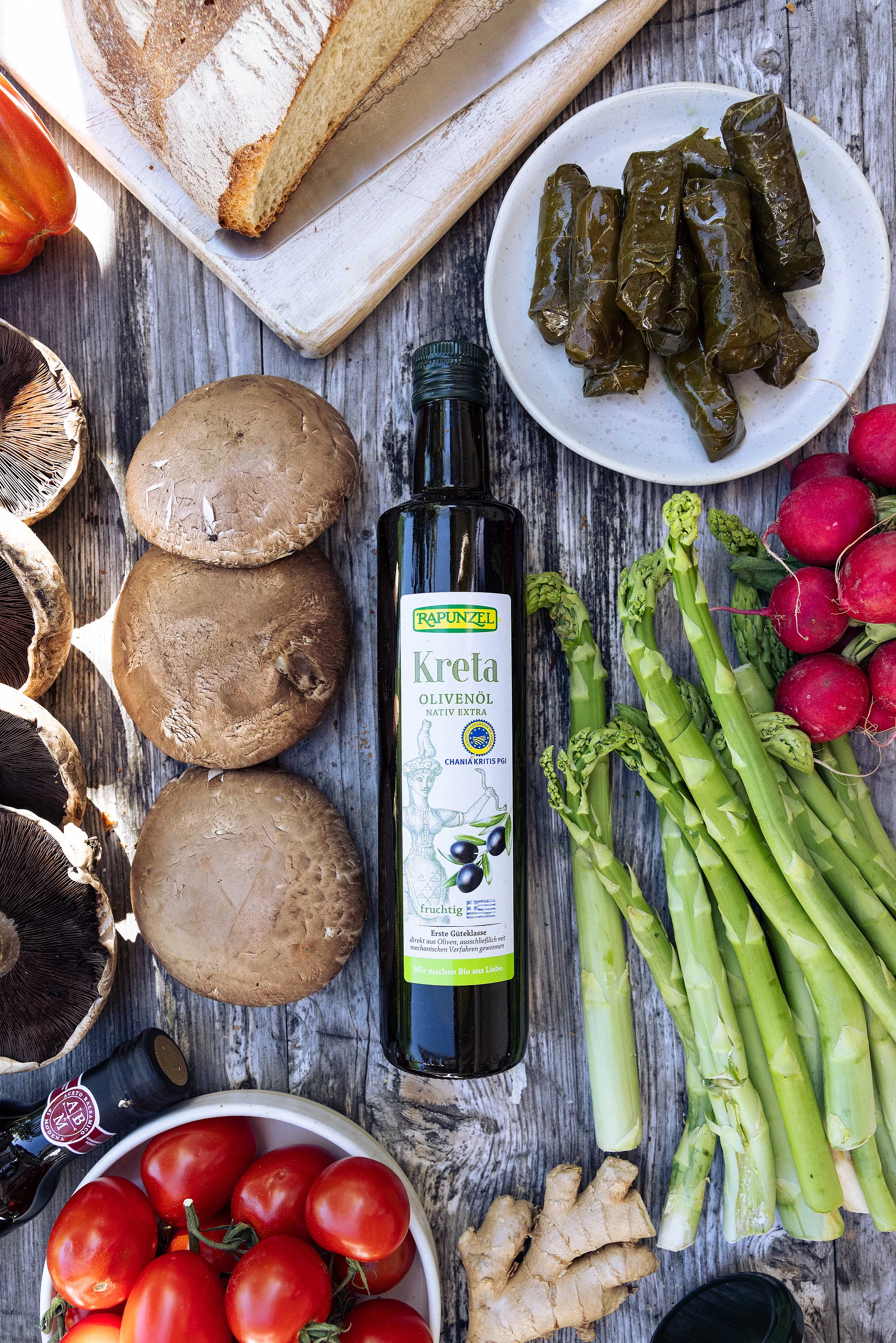 Rapunzel Olivenöl Kreta – Bio-Qualität für mediterrane Küche. Flasche Rapunzel Olivenöl aus Kreta auf Tisch mit grünem Spargel, Pilzen und Weinblättern – vielseitig für mediterrane Gerichte.