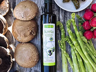 Rapunzel Olivenöl Kreta – Bio-Qualität für mediterrane Küche. Flasche Rapunzel Olivenöl aus Kreta auf Tisch mit grünem Spargel, Pilzen und Weinblättern – vielseitig für mediterrane Gerichte.