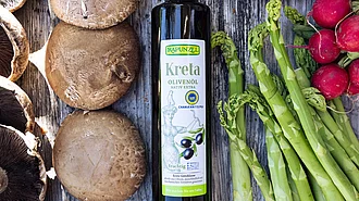 Flasche Rapunzel Olivenöl aus Kreta auf Tisch mit grünem Spargel, Pilzen und Weinblättern – vielseitig für mediterrane Gerichte.