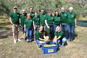 Rapunzel Mitarbeiterinnen und Mitarbeiter helfen 2024 bei der Olivenernte auf Kreta, vor ihnen eine blaue Kiste mit frisch geernteten Bio-Oliven.