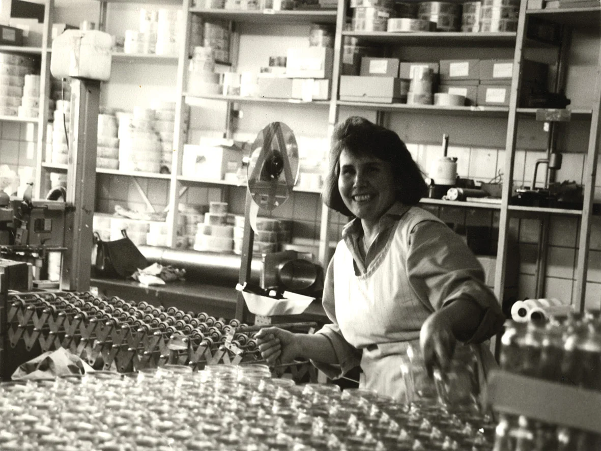 Mitarbeiterin bei der Abfüllung von Nussmus in den 1970er-Jahren. Historisches schwarz-weiß Foto zeigt eine Mitarbeiterin vor einer Lage leerer Gläser für die Nussmuss-Abfüllung.