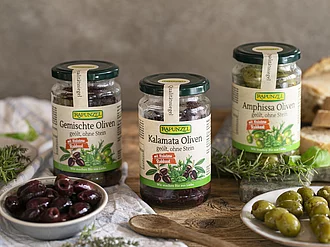 Drei Gläser gefüllt mit Oliven, davor Kalamata und Amphissa Oliven in Schälchen.