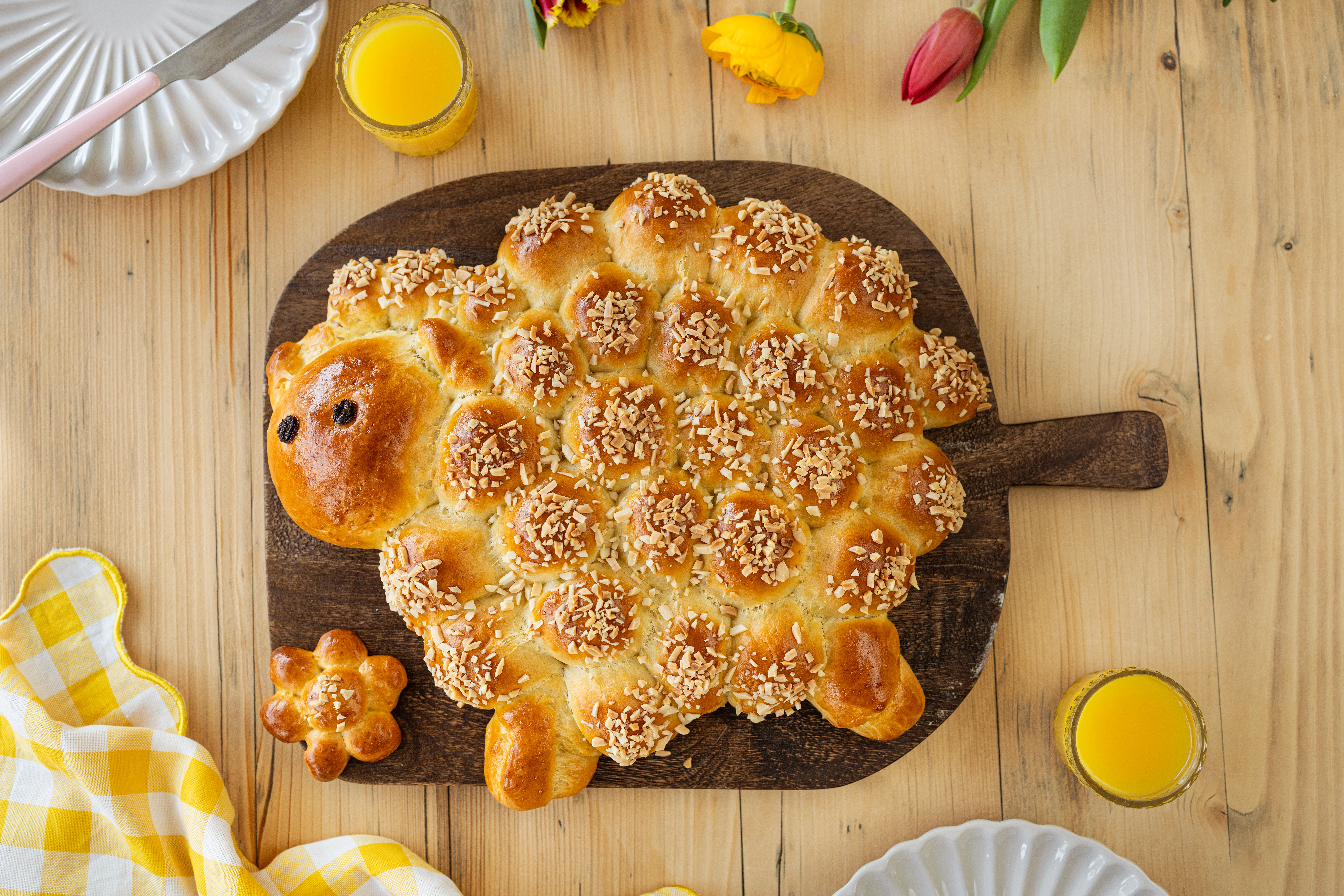 Osterlamm aus Hefeteig mit Mandelstiften auf einem dunklen Holzbrett, umgeben von Frühstücksgedeck und Frühlingsblumen