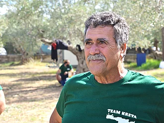 Dimitri Katzaros Auf dem Bild ist unser kretischer Oliven-Lieferant Dimitri vor einem verschwommenen Olivenbuam zu sehen. Er schaut leicht an der Kamera vorbei und freut sich
