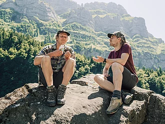 Zwei Wanderer haben sich zur Rast auf einem Großen Stein gesetzt, einer von ihnen hält eine Tüte salzige Snacks von Rapunzel in den Händen.