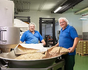 Zwei Mitarbeiter füllen Rohware aus Säcken in den Müsli-Mischer.