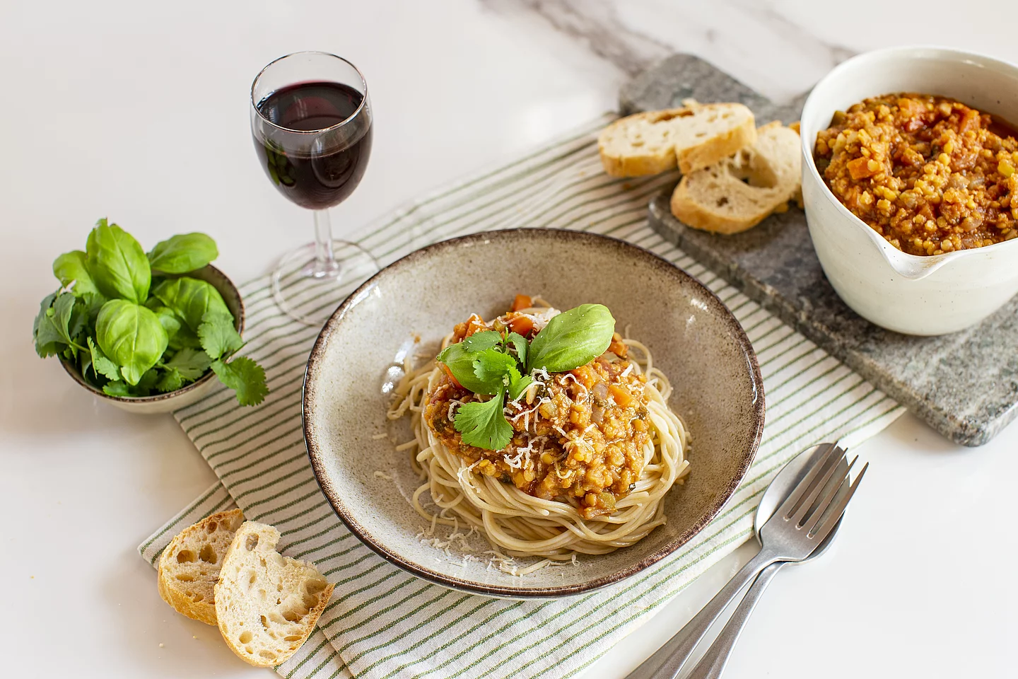 Ein tiefer Keramikteller mit Spaghetti und einer herzhaften Linsen-Bolognese, garniert mit frischem Basilikum und Koriander. Im Hintergrund stehen eine Schüssel mit der restlichen Sauce aufgeschnittenes Baguette, ein Glas Rotwein und eine kleine Schale mit Kräutern auf einer hellen Tischoberfläche