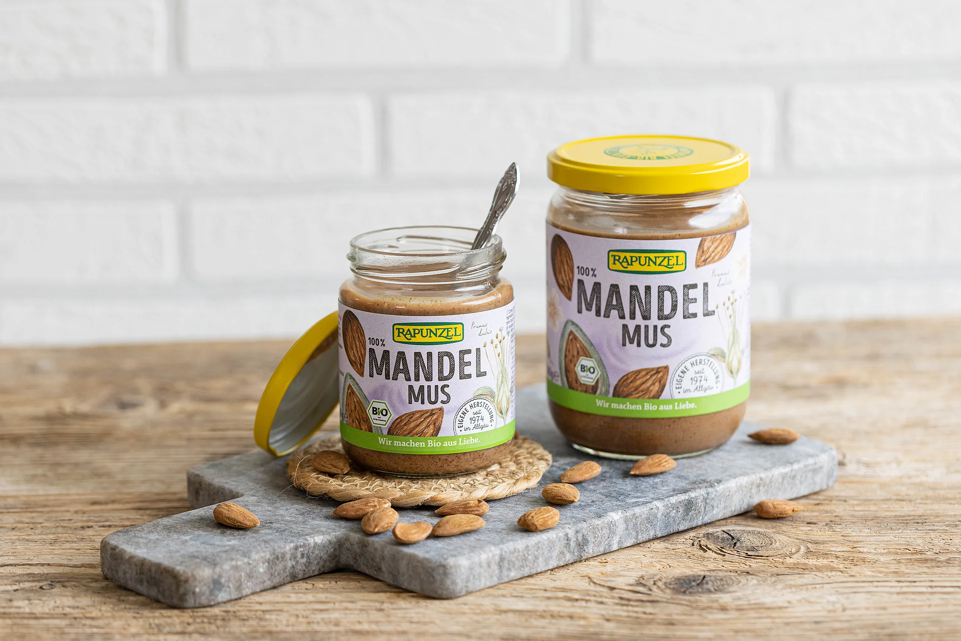 Verführerisches Mandelmus – ein Rapunzel Klassiker von Beginn an. Zwei Mandelmuse braun in zwei verschiedenen Größen stehen auf einer Servierplatte aus Stein auf einem Holztisch.