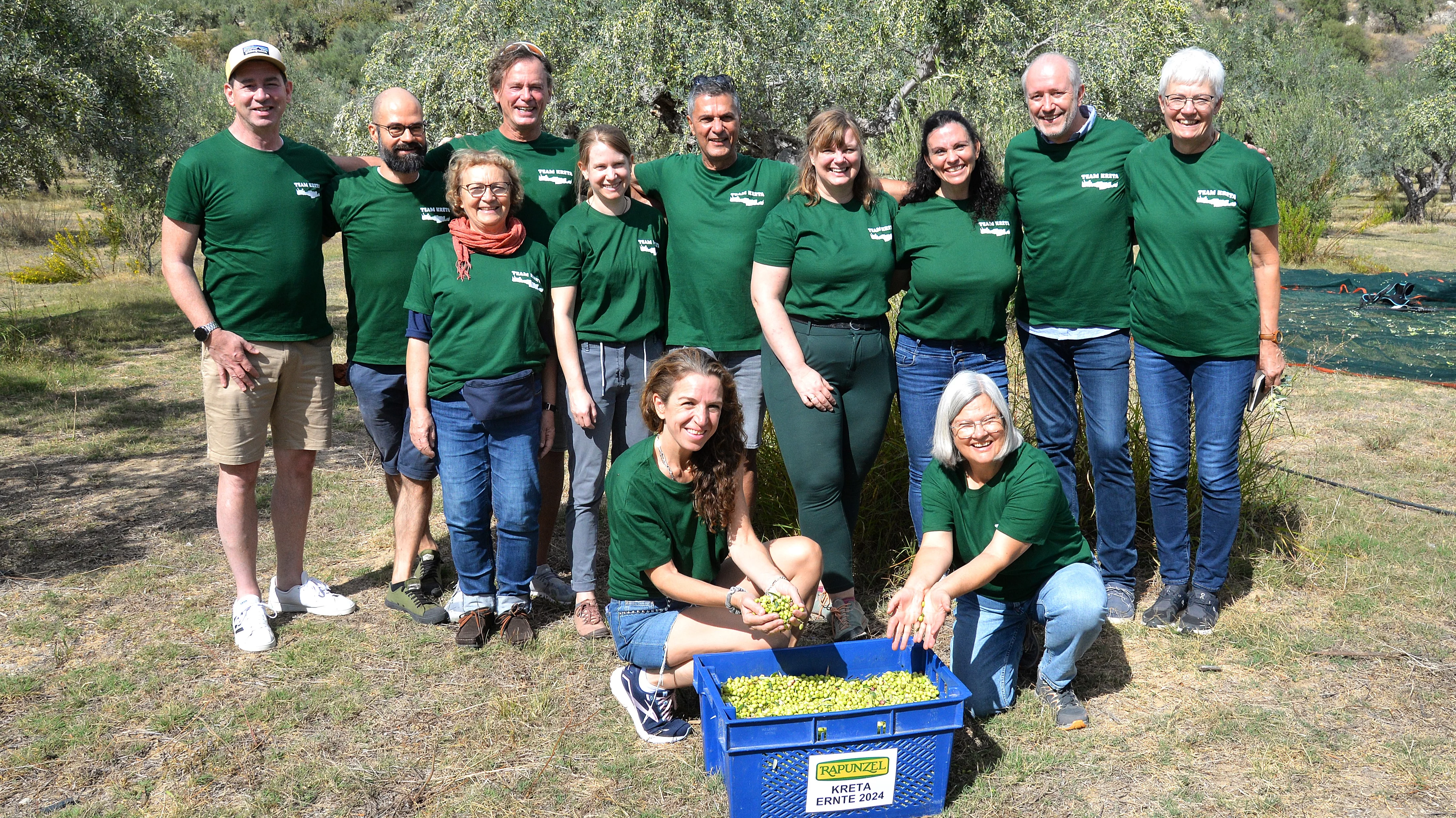 Rapunzel Mitarbeiterinnen und Mitarbeiter helfen 2024 bei der Olivenernte auf Kreta, vor ihnen eine blaue Kiste mit frisch geernteten Bio-Oliven.
