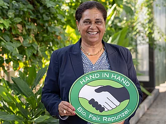 Firmengründerin Samia Chibani mit einem HAND IN HAND-Schild im Tropenhaus bei Rapunzel in Legau. 