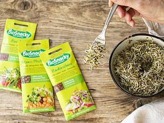 Drei Packungen Keimsaaten von Rapunzel bioSnacky neben Schale mit frischen Sprossen.