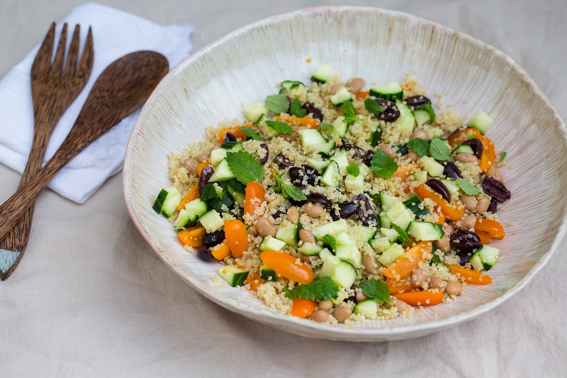 Quinoa-Salat mit Borlotti-Bohnen, Oliven und Minz-Blättern. Quinoa-Salat mit Borlotti-Bohnen, Oliven und Minz-Blättern, angerichtet auf einem Teller, der auf einem Tischtuch steht, daneben Holzbesteck und eine Serviette.