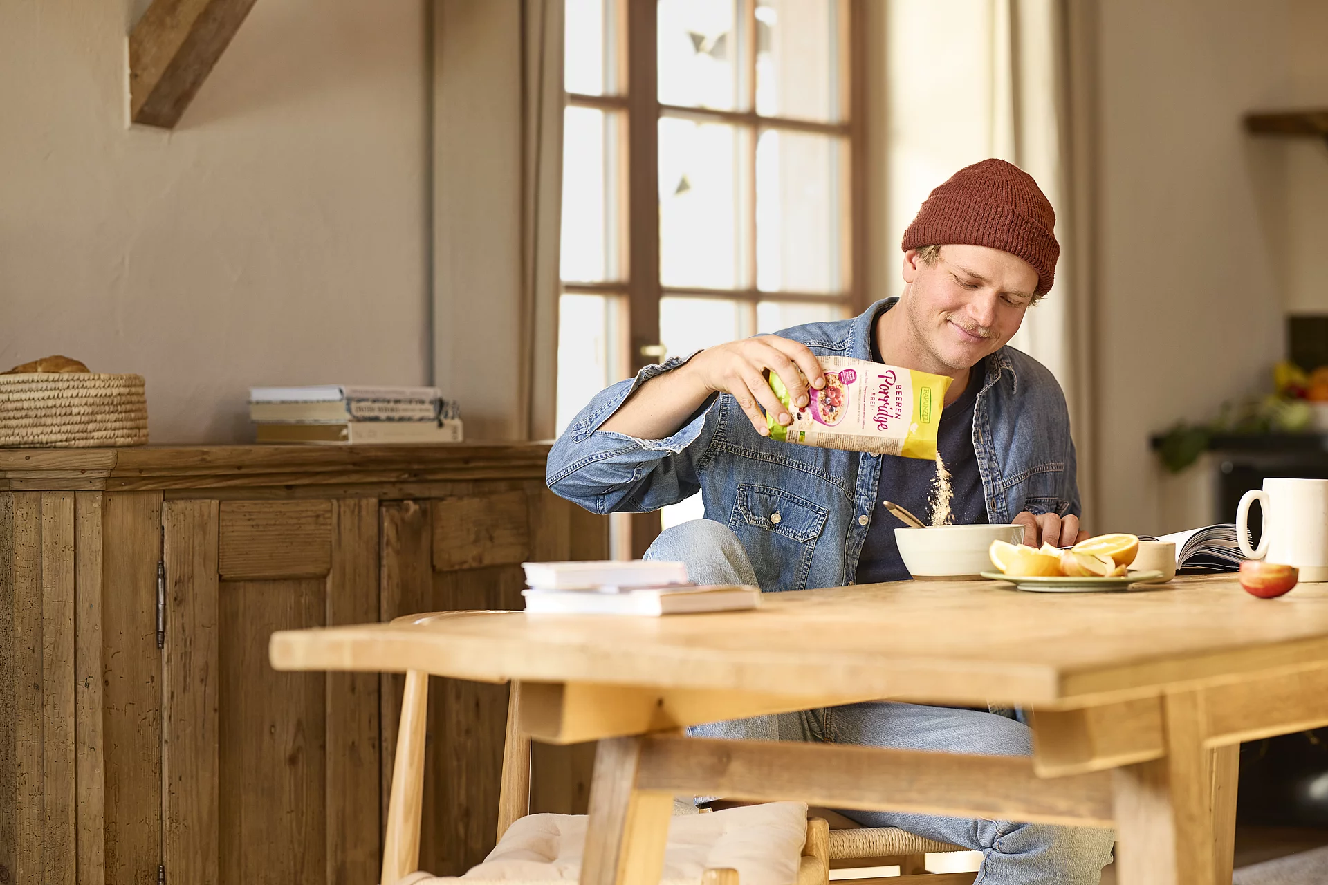 Porridge für einen entspannten Start in den Tag. Ein junger Mann sitzt in einer gemütlichen Bauernhofküche und füllt sich Porridge in eine Müslischüssel. Auf dem Tisch liegen Zeitschriften und eine Schale mit.