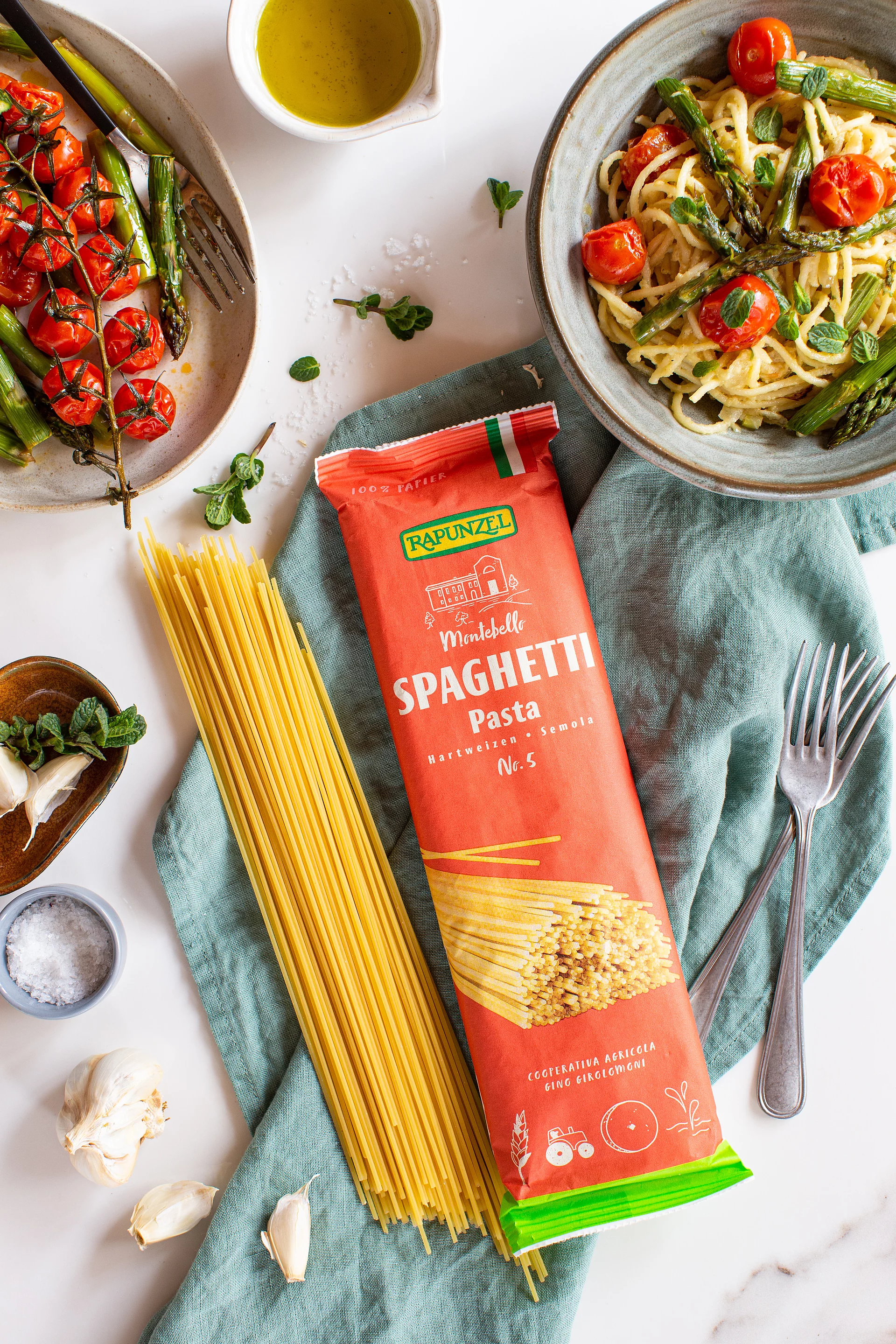 Rapunzel Bio-Spaghetti werden sowohl in der Originalverpackung als auch lose auf einem Küchentuch präsentiert. Drumherum ist ein angerichtetes Nudelgericht mit Gemüse dekorativ angerichtet.