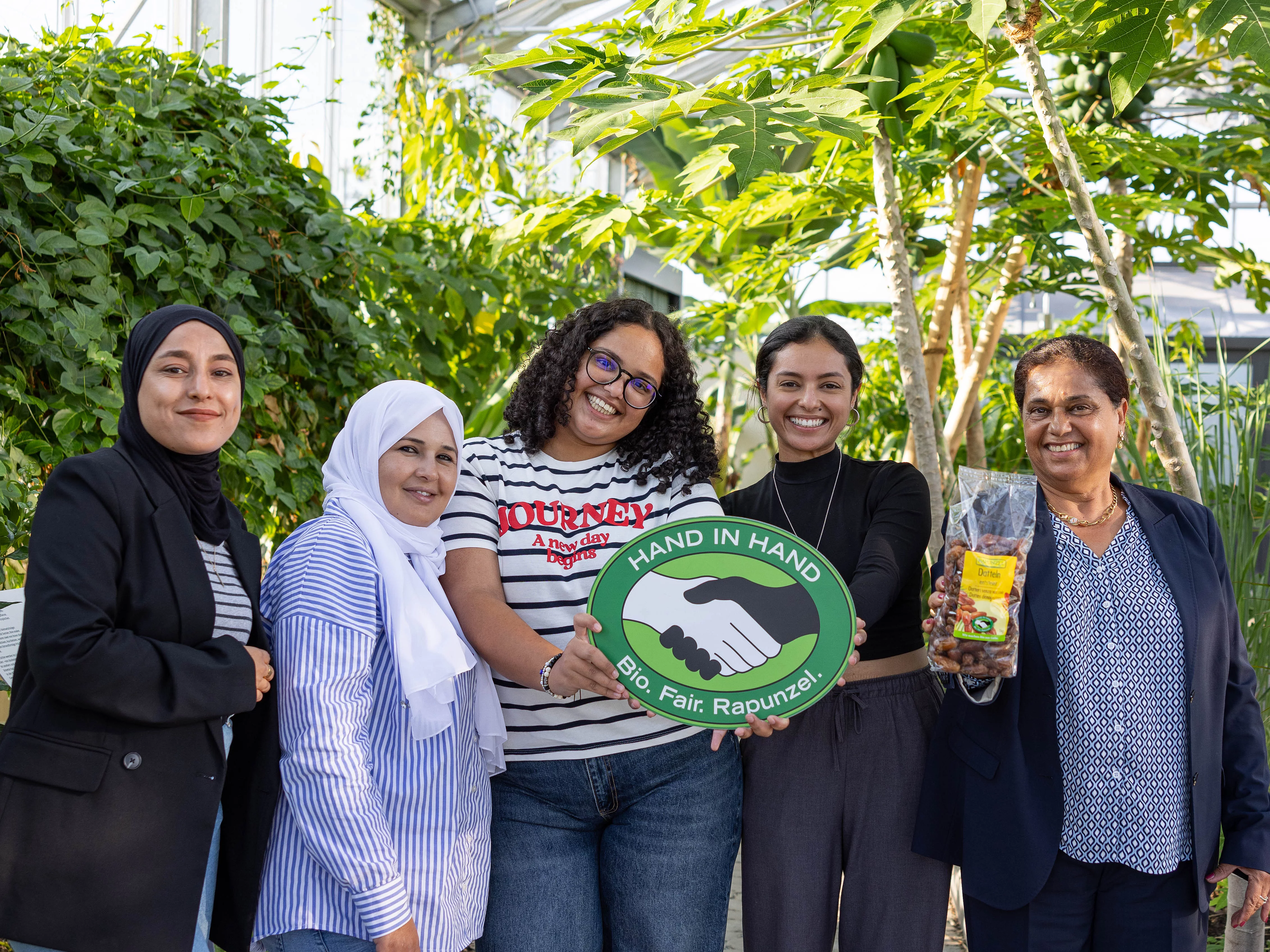 Gruppenfoto im Tropenhaus von Rapunzel mit CCF-Gründerin Samia Chibani. Mit im Bild sind das HAND IN HAND-Logo und die Datteln, die von Rapunzel verkauft werden.