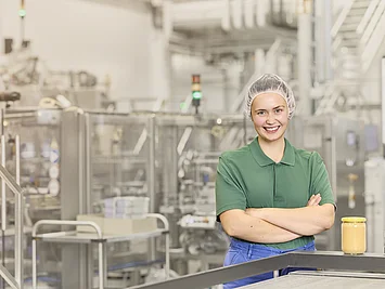 Mitarbeiterin mit Haarnetz vor einer Nussmus-Anlage, davor ein fertig abgefülltes Glas ohne Etikett