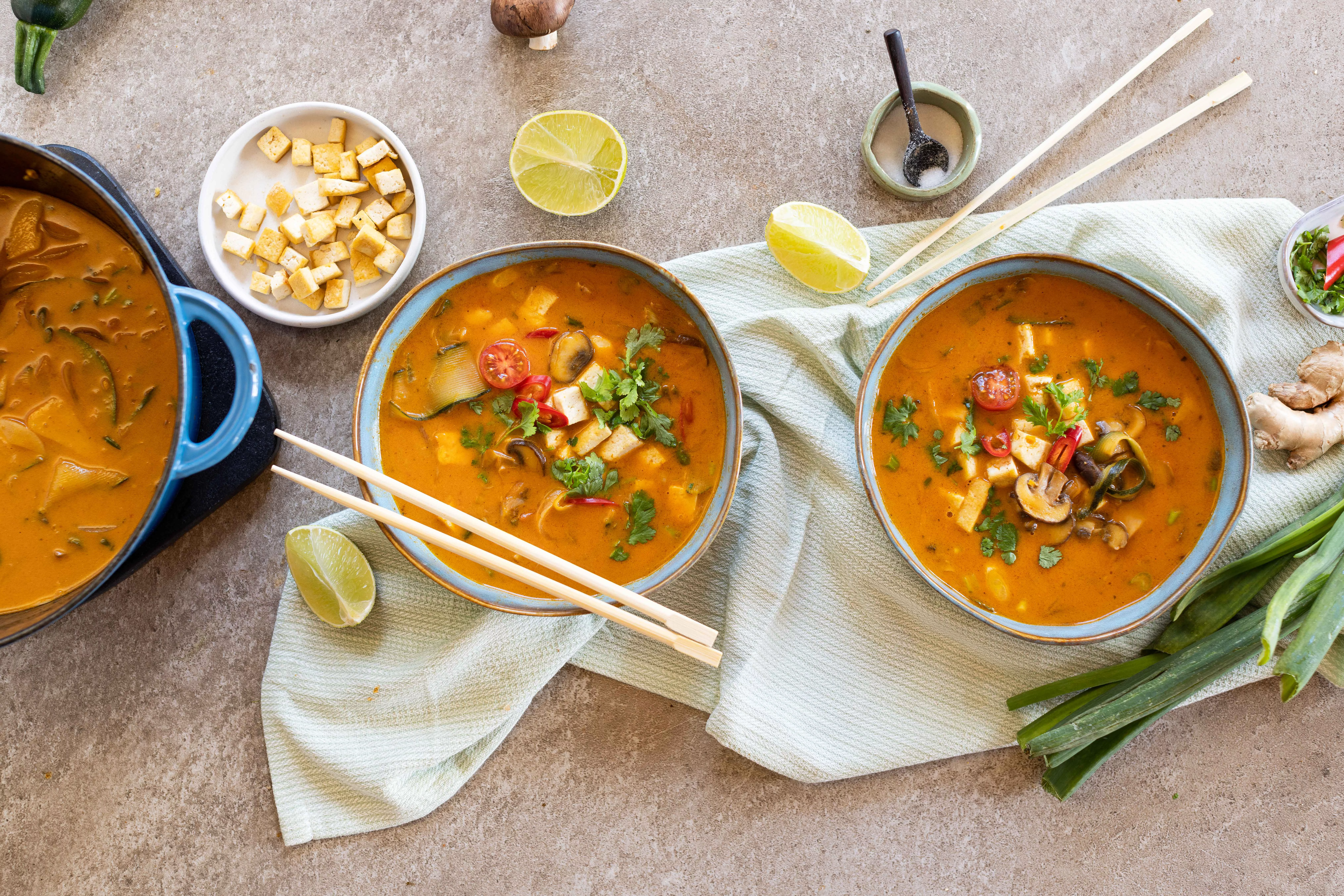 Rote Tom-Yum-Suppe in heller Schale mit Tofuwürfeln, Pilzen, Paprikastreifen, frischen Kräutern und Limettenspalten.