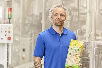 Mitarbeiter aus der Produktion steht neben der Abpackmaschine und präsentiert fertig abgepacktes Müsli.