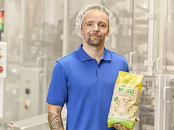 Mitarbeiter aus der Produktion steht neben der Abpackmaschine und präsentiert fertig abgepacktes Müsli.