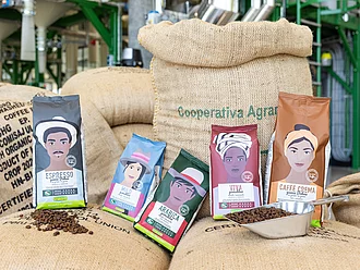 Eine Auswahl von Rapunzel Heldenkaffees vor einem Kaffeesack im Produktionsumfeld