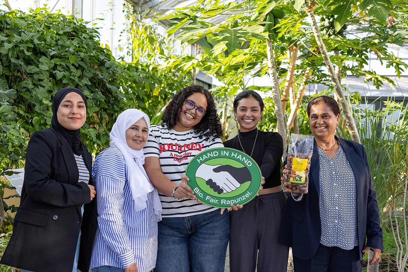 Gruppenfoto im Tropenhaus von Rapunzel mit CCF-Gründerin Samia Chibani. Mit im Bild sind das HAND IN HAND-Logo und die Datteln, die von Rapunzel verkauft werden.