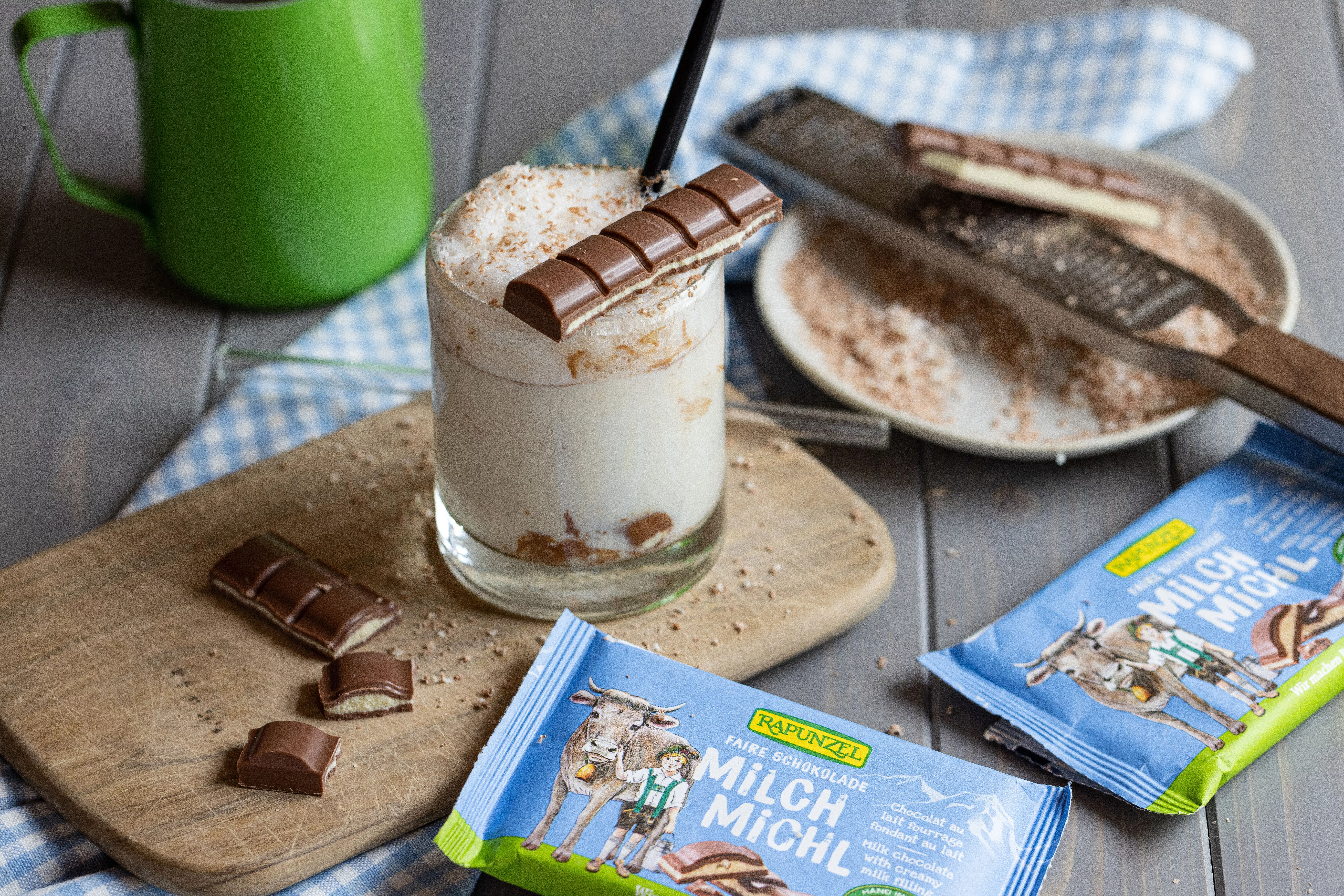Eine Tafel Rapunzel Milch-Michl Schokolade liegt im Vordergrund, dahinter steht ein Glas mit einem Getränk, leicht unscharf im Hintergrund