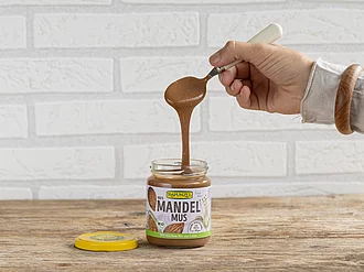 Glas Rapunzel Mandelmus und Mandelmus braun, stimmungsvolle Küchenaufnahme mit Hand und Löffel – natürliche Bio-Qualität für vielseitigen Genuss.