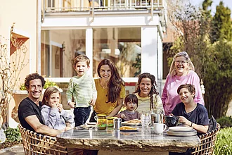 Familie Wilhelm am Tisch im Garten. Familie Wilhelm sitzt im Garten an einem Tisch, manche Familienmitglieder stehen auch, und es stehen verschiedene Rapunzel Produkte auf dem Tisch.