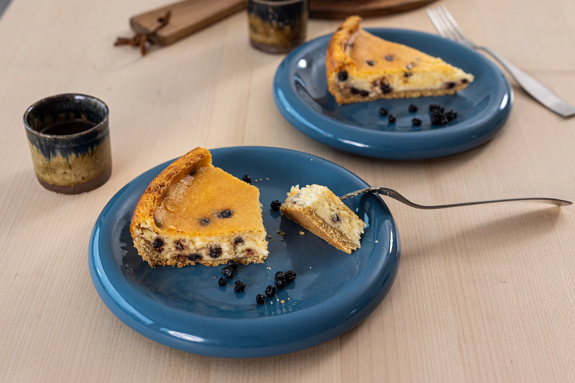 Käsekuchenstücke mit Blaubeeren auf zwei Tellern, daneben Becher mit Kaffee.