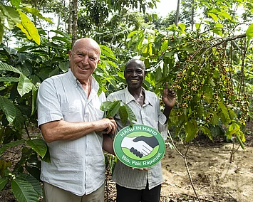 Joseph Wilhelm neben einem Kaffeebauern auf seiner Parzelle. Gemeinsam halten sie das HAND IN HAND-Logo.