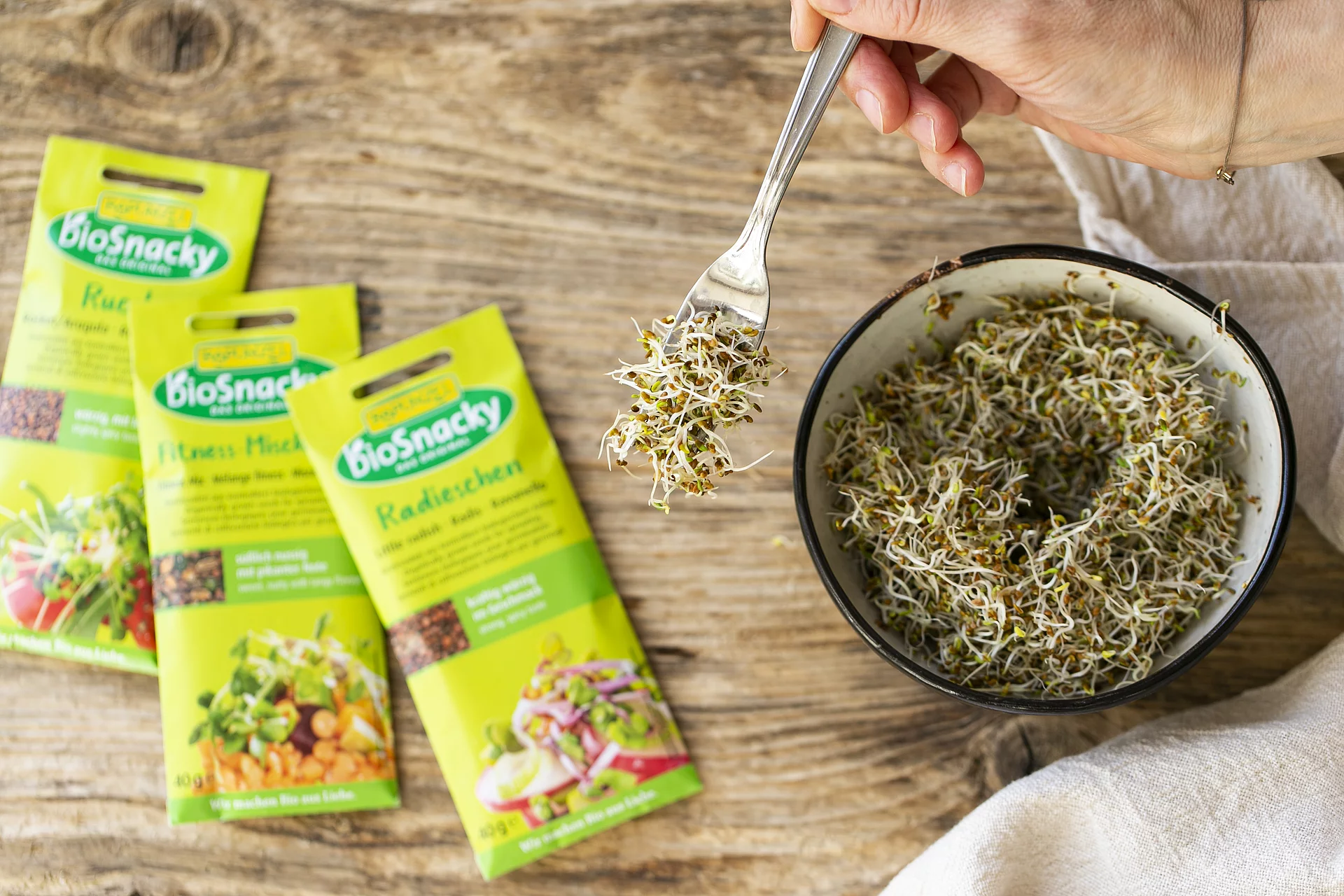 Verschiedene Keimsaaten von Rapunzel bioSnacky. Drei Packungen Keimsaaten von Rapunzel bioSnacky neben Schale mit frischen Sprossen.