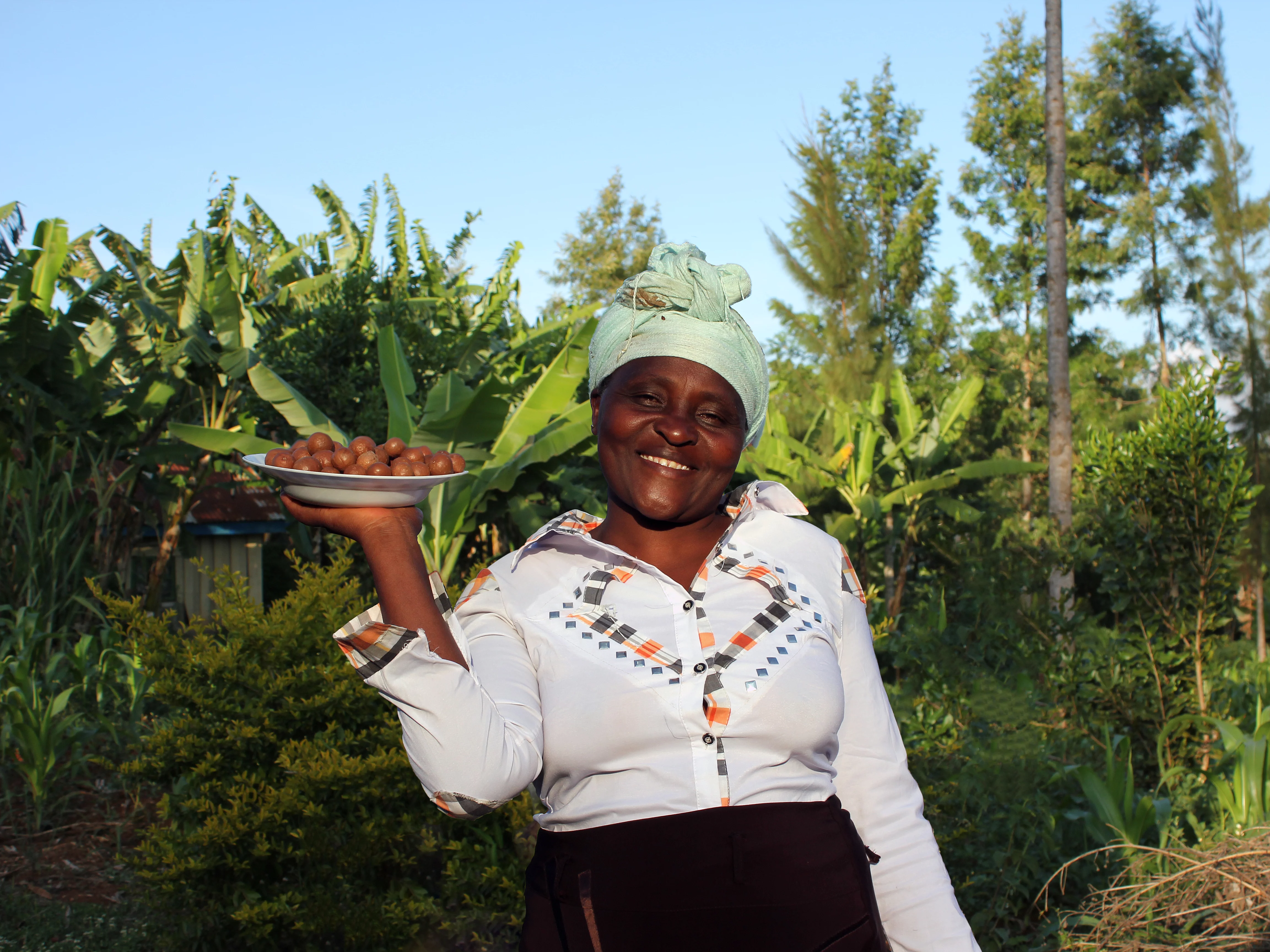 Eine kenianische Bäuerin die Macadamias auf einem Teller präsentiert und auf dem Feld steht. 