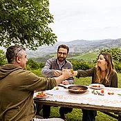 Drei Geschwister sitzen miteinander an einem Tisch und essen Pasta, dahinter die italienischen Marken.