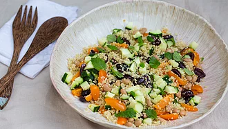 Quinoa-Salat mit Borlotti-Bohnen, Oliven und Minz-Blättern, angerichtet auf einem Teller, der auf einem Tischtuch steht, daneben Holzbesteck und eine Serviette.