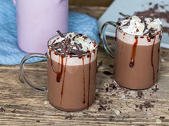 Auf einem Holztisch stehen zwei Tassen heiße Schokolade mit Sahne und Schokoladensauce garniert.Im Hintergrund steht, leicht angeschnitten, Küchengeschirr