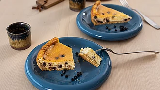 Käsekuchenstücke mit Blaubeeren auf zwei Tellern, daneben Becher mit Kaffee.