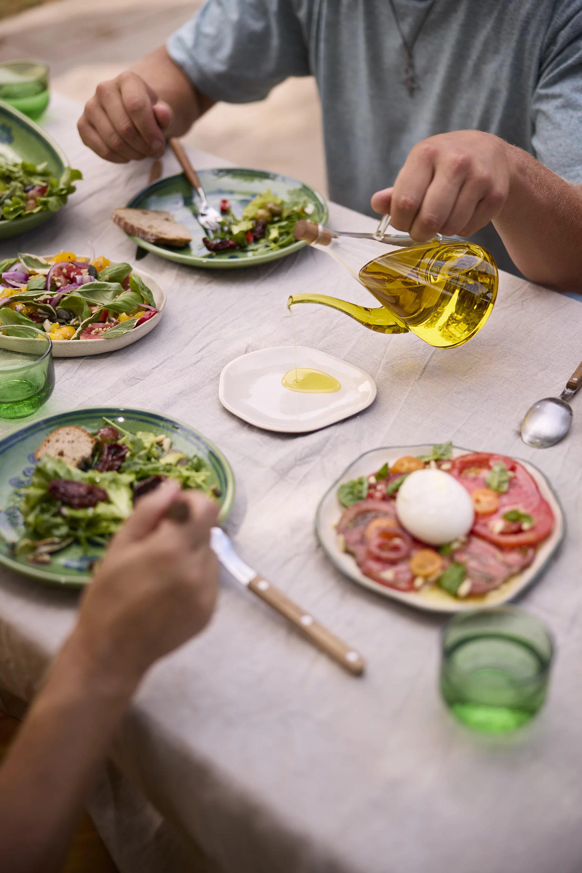 Mediterrane Salatvariationen mit Rapunzel Bio-Olivenöl. Tisch mit frischen Salatvariationen, Tomaten-Mozzarella und Olivenöl-Dekantierer – Rapunzel Bio-Olivenöl für mediterrane Küche
