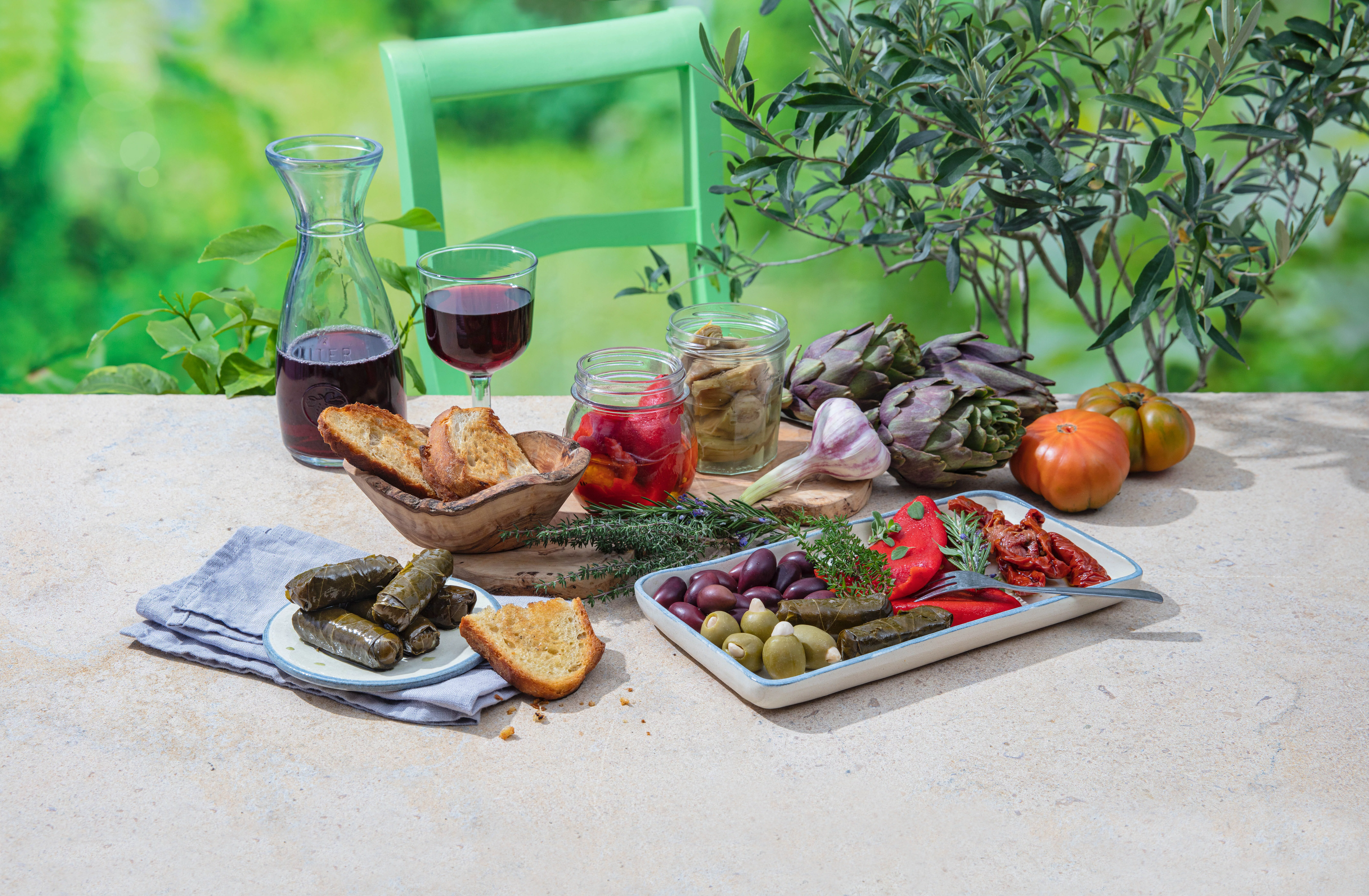 Gedeckter Tisch mit bunter Antipasti-Auswahl wie Dolmas, Oliven, Paprika, Tomaten, Artischocken, dazu Rotwein.