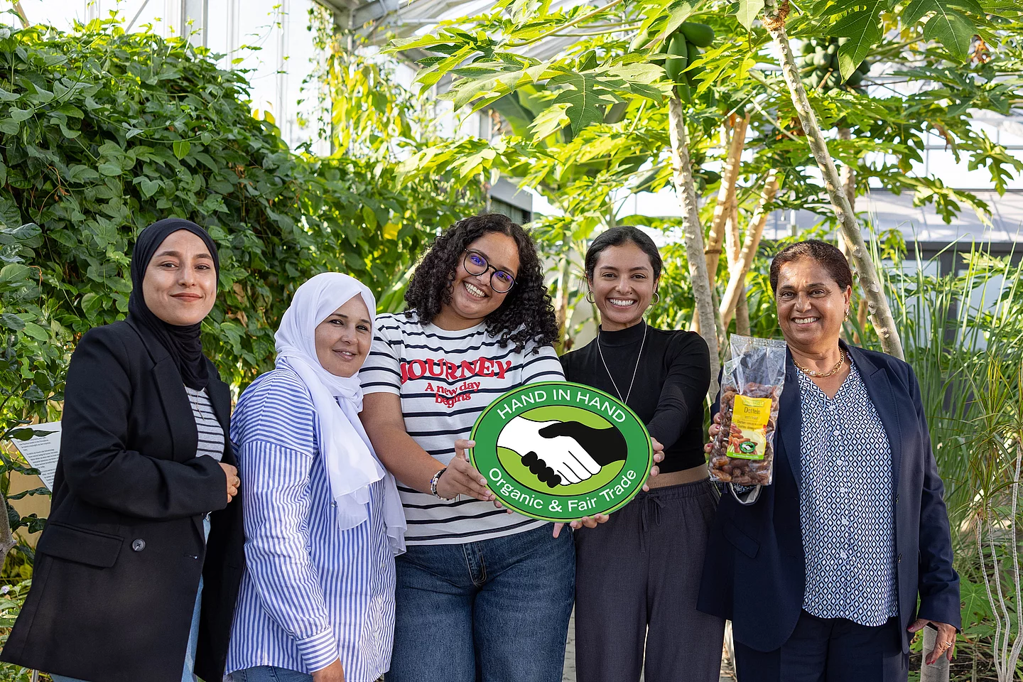 CCF zu Besuch bei Rapunzel in Legau. Gruppenfoto im Tropenhaus von Rapunzel mit CCF-Gründerin Samia Chibani. Mit im Bild sind das HAND IN HAND-Logo und die Datteln, die von Rapunzel verkauft werden.