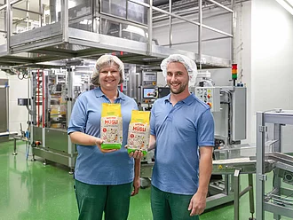 Rapunzel Produktionsmitarbeiter stellen Müsli her. Zwei Mitarbeiter aus der Rapunzel Produktion halten je eine Packung Müsli in die Kamera, im Hintergrund die Müslianlage.