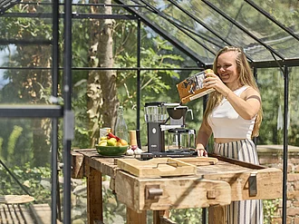 Kaffeezubereitung mit Rapunzel Kaffee Sommerliche Szene in einem Gewächshaus aus Glas - Eine junge Frau gibt Kaffeepulver in den Filter einer Kaffeemaschine.
