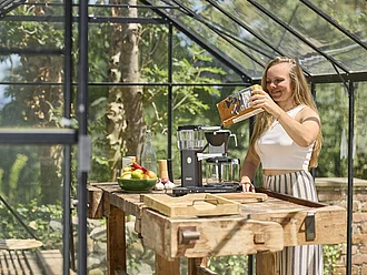 Sommerliche Szene in einem Gewächshaus aus Glas - Eine junge Frau gibt Kaffeepulver in den Filter einer Kaffeemaschine.