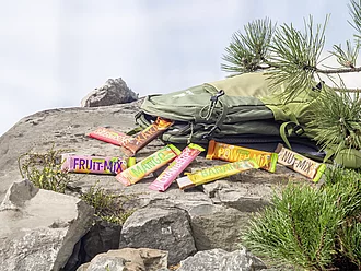 Auf einem Felsen liegt ein Wanderrucksack, aus dem Rapunzel Fruchtschnitten herausgefallen sind.