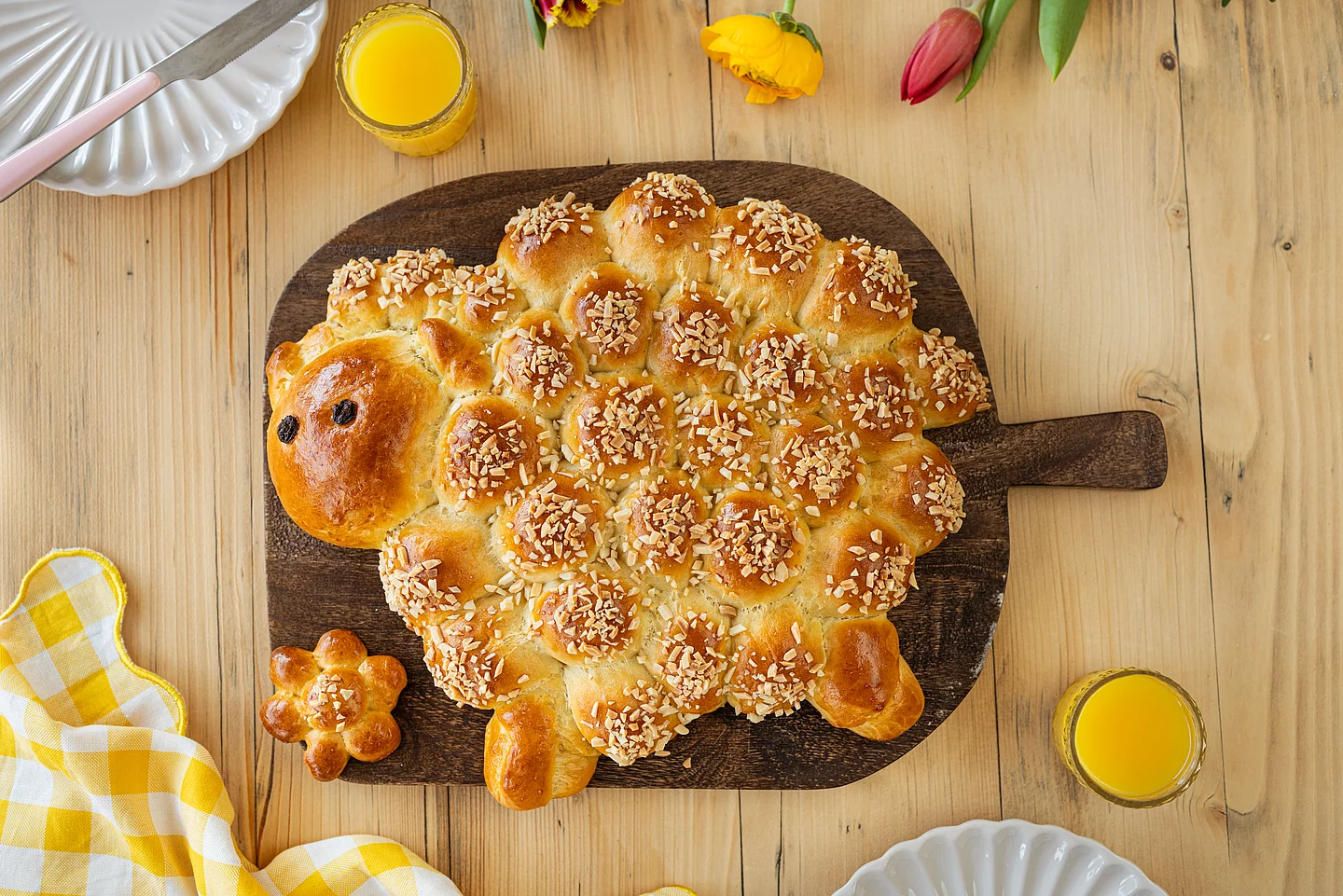 Osterlamm aus Hefeteig Osterlamm aus Hefeteig mit Mandelstiften auf einem dunklen Holzbrett, umgeben von Frühstücksgedeck und Frühlingsblumen