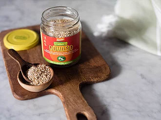 Ein Glas Gomasio mit offenem Deckel und daneben ein Löffel mit Gomasio auf einem Holzbrett.