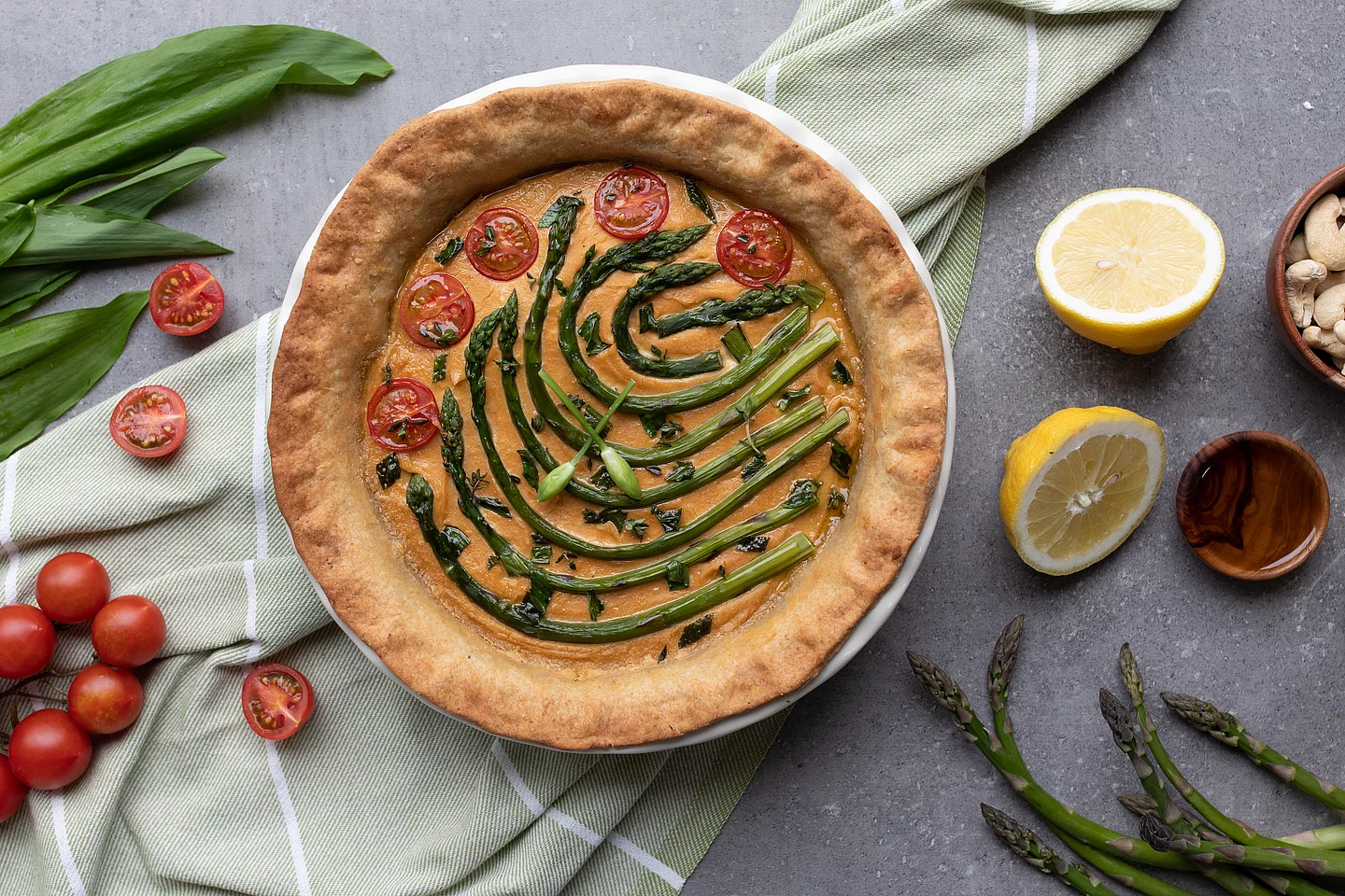 Spargel-Bärlauch-Tomaten-Tarte mit Nussboden. Draufsicht auf eine runde, hausgemachte Spargel-Tarte mit einem dunklen Nussboden und knusprigem Rand. Sie ist mit ganzen grünen Spargelstangen, roten Tomatenhälften und frischem Bärlauch belegt. Das Gericht liegt auf einem hellen Teller auf einer dunklen, neutralen Arbeitsplatte. Ringsum sind die verschiedenen Zutaten der Tart dekorativ platziert.
